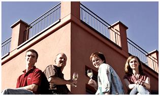 Vier lachende Menschen sitzend mit Weingläsern vor orangen Haus